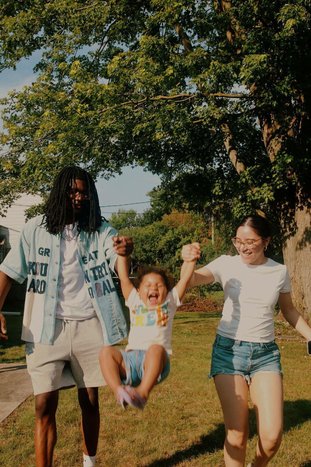 A group of people in white shirts and denim shorts run playfully through a sunny field with trees in the background.