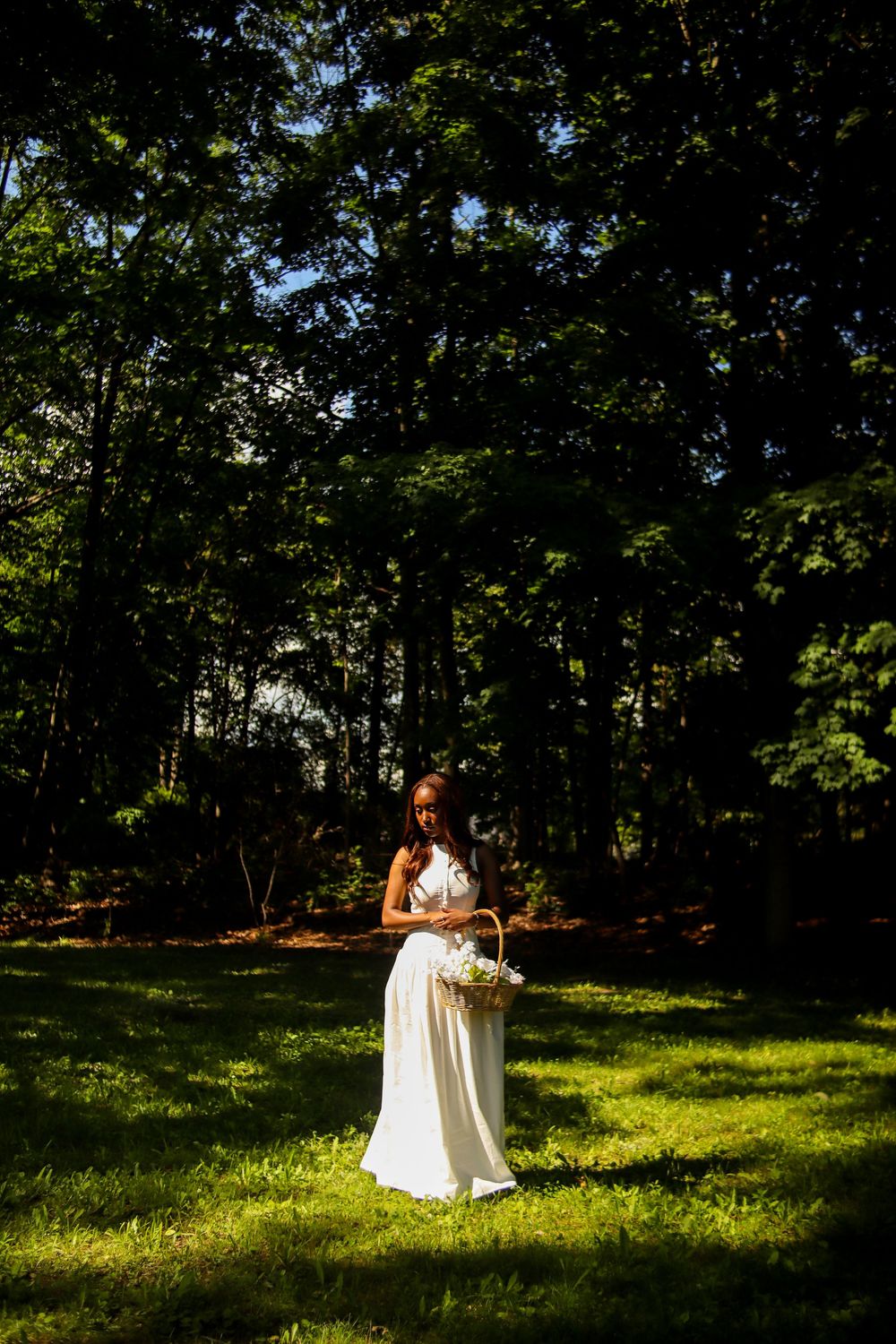 Bride in white gown holding flower basket stands in sunlit forest clearing surrounded by tall green trees.