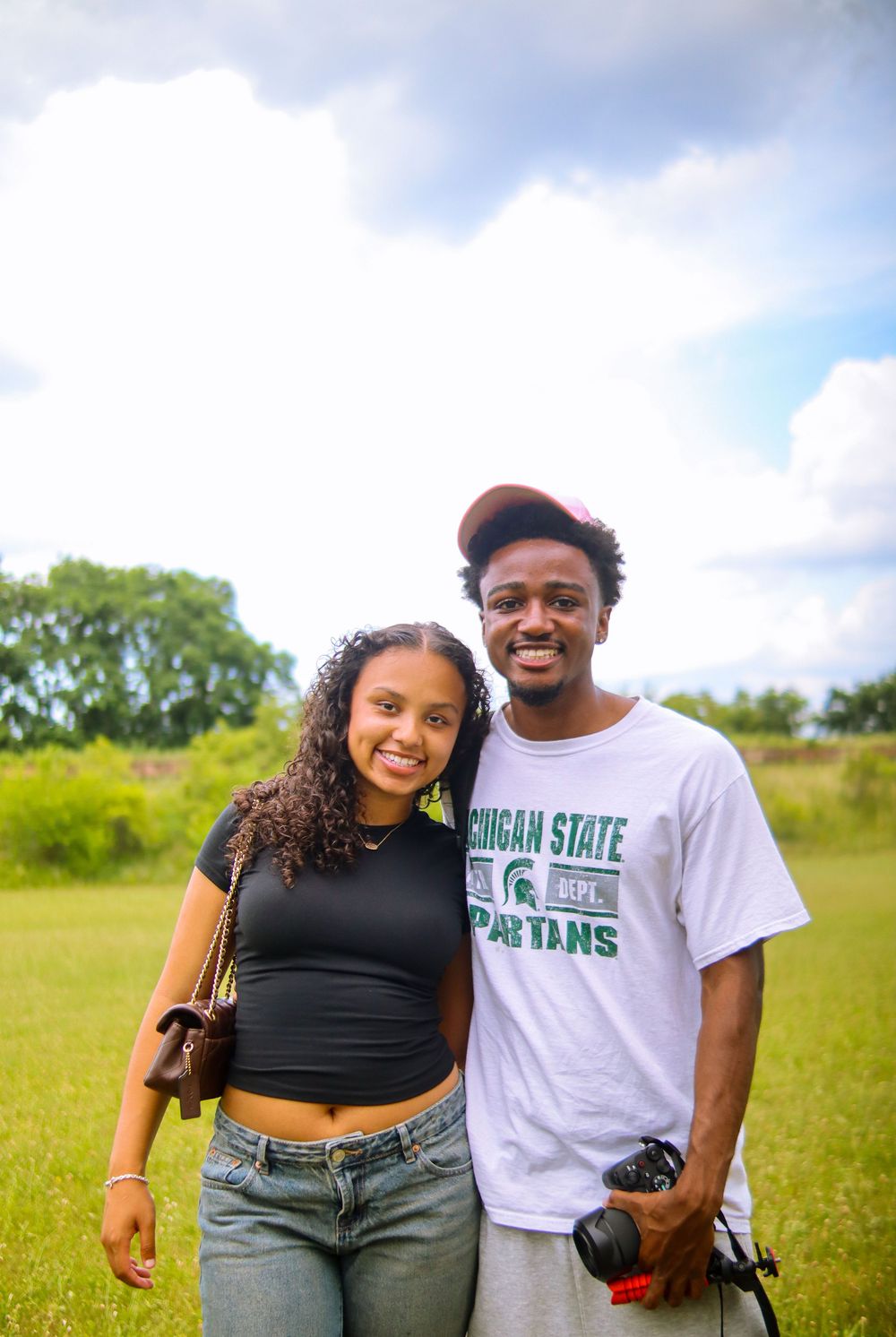 A young couple smiles outdoors on a grassy field, man wearing Michigan State Spartans shirt holding a camera.