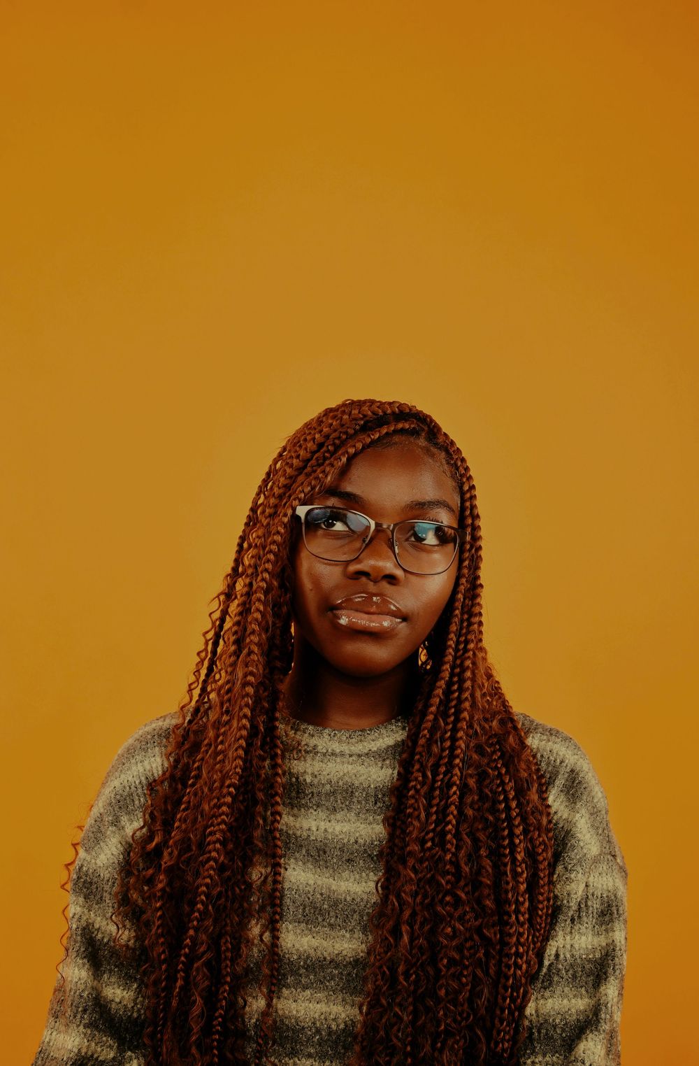 A person with glasses and long reddish-brown box braids smiles warmly while wearing a gray sweater against an orange background.