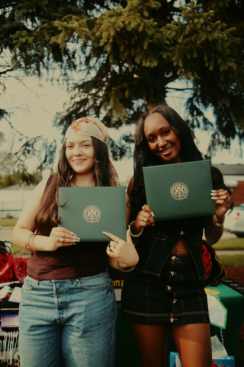 Two graduates proudly display their green diplomas while standing outdoors on a sunny day near evergreen trees.