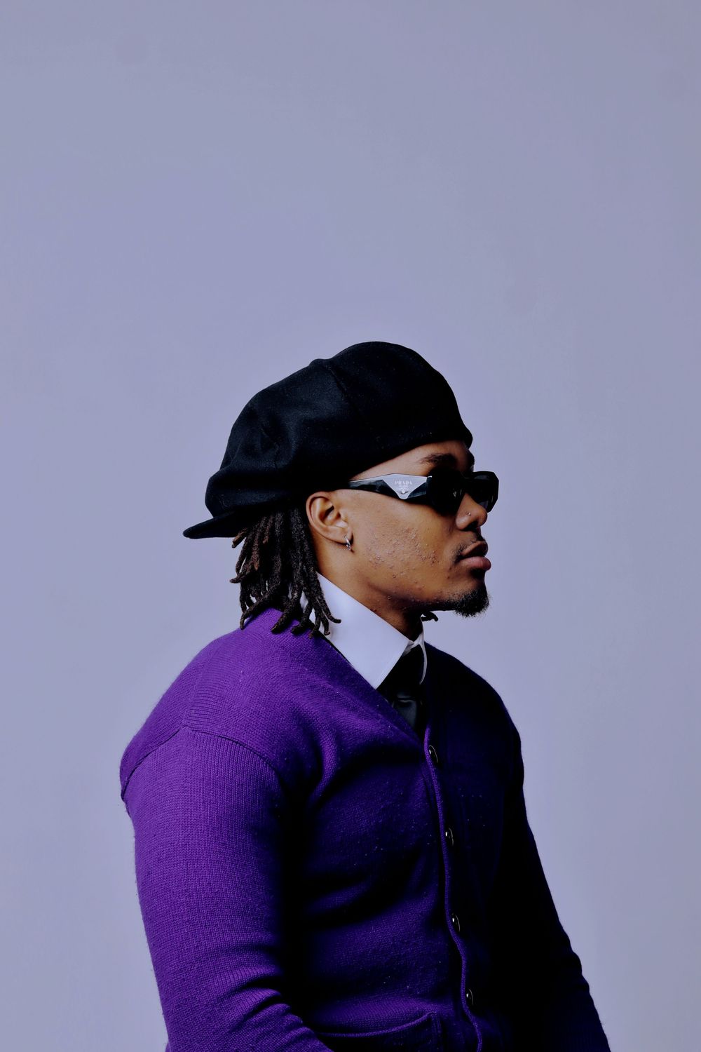 A person in a purple sweater and black baseball cap poses against a gray background in a profile view.