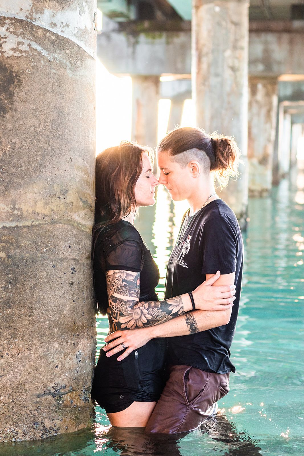 Couple embraces romantically in blue water between concrete pillars during sunset photoshoot under lake worth pier
