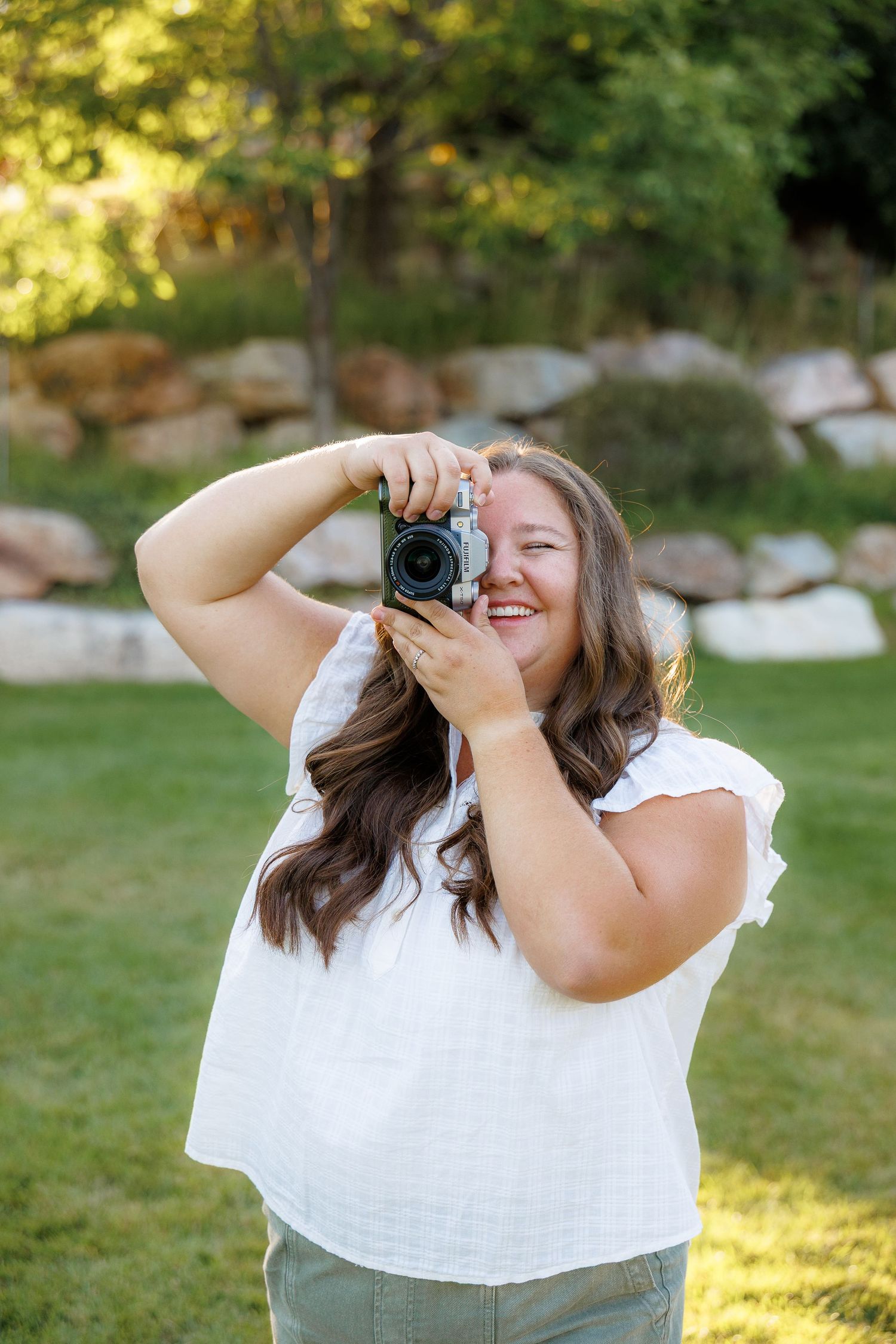 Smiling woman in white top holding a camera outdoors in a green park setting with rocks and trees.