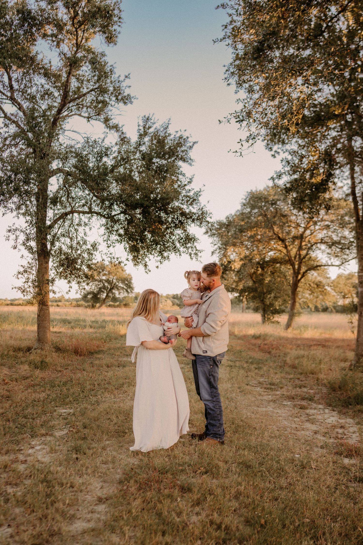 A couple stands together in a peaceful rural setting with oak trees at golden hour in a grassy field.