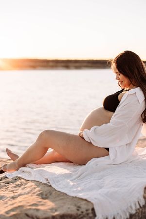 Pregnant woman with long dark hair wearing bikini and white shirt on sand near water