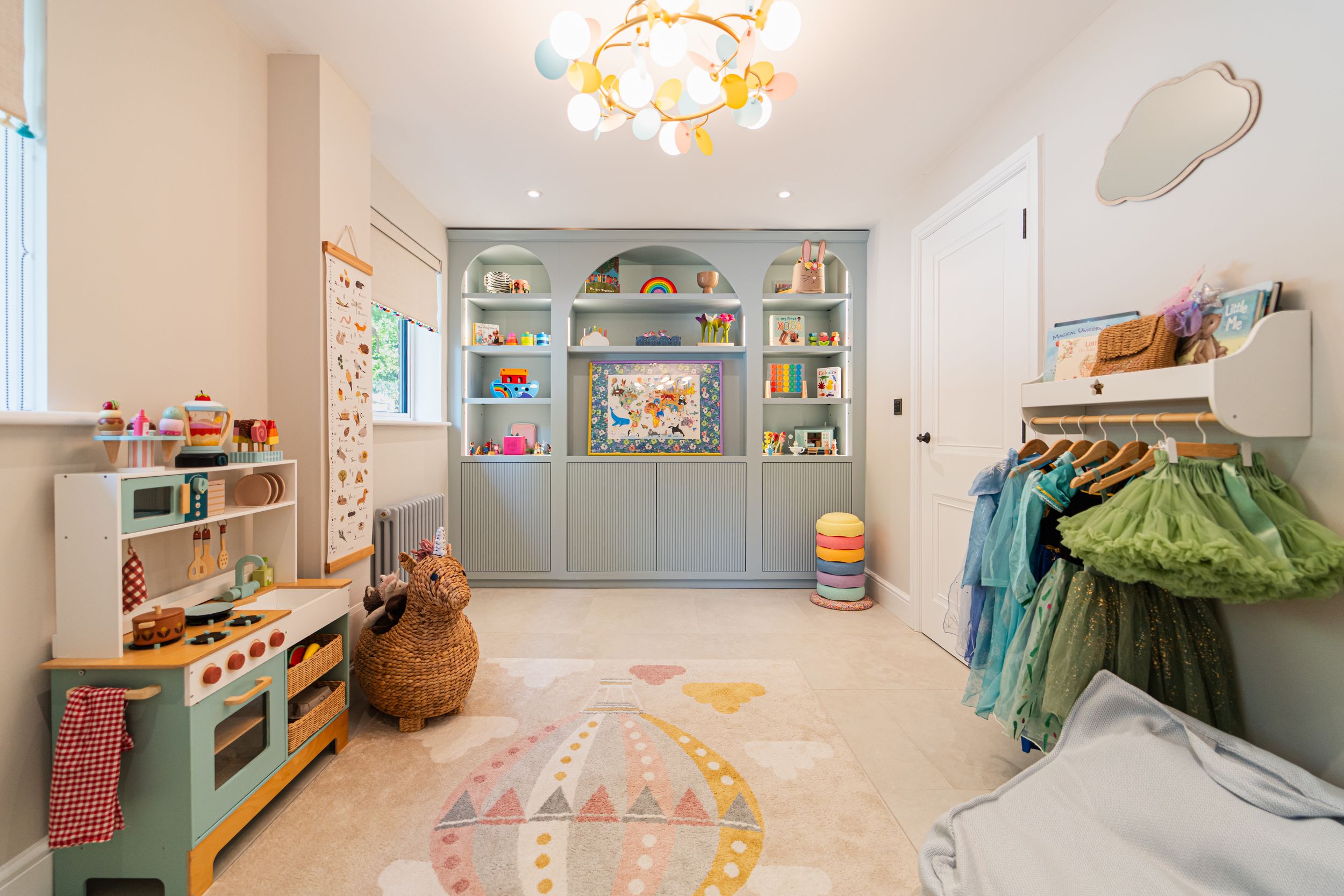 A bright playroom with built-in shelving, a play kitchen, and a round pastel rug features organized storage and hanging space.