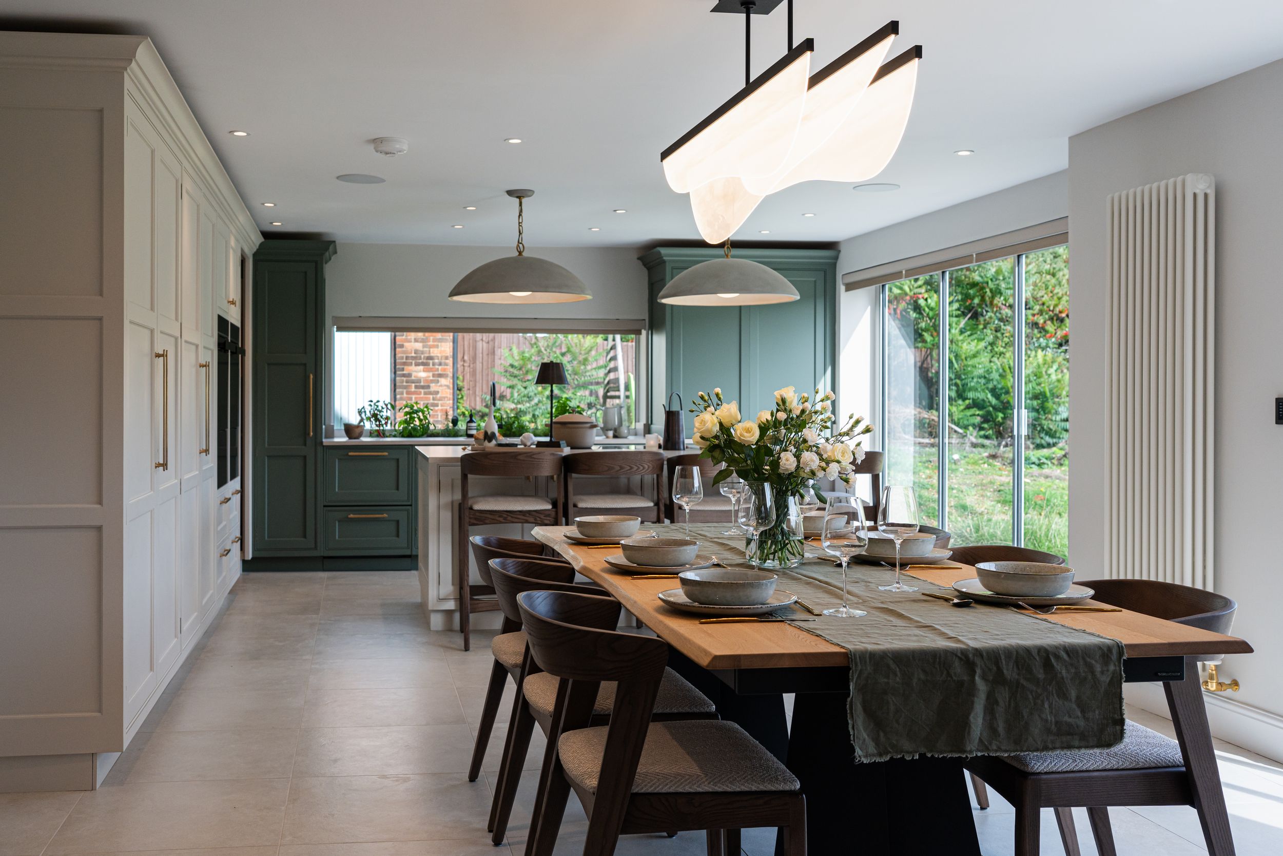 Modern open concept kitchen and dining area with dark green cabinets and wooden dining table illuminated by pendant lights.