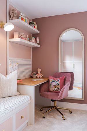 Pink home office nook with built-in desk, floating shelves, arched window, and velvet swivel chair.