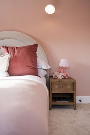 Cozy bedroom with pink walls featuring a bed with mauve pillows and wooden nightstand with rattan detailing.