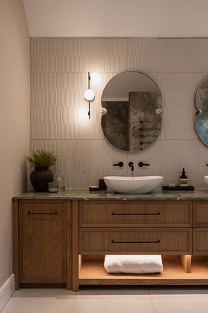 Modern bathroom vanity with floating wooden cabinets, vessel sink, and oval mirror against textured white tile wall.