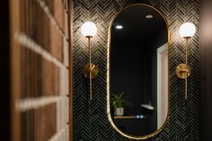 Contemporary bathroom featuring dark herringbone tile walls and round gold-framed mirror with accent lighting.