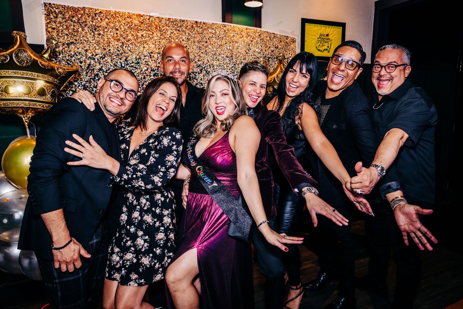 Group of party guests posing playfully together at a social gathering with decorative gold and patterned wallpaper background.