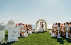Scenic outdoor wedding ceremony on a grassy hilltop with white floral arch and seated guests against blue sky backdrop.