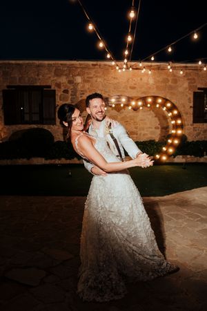 Newlyweds dance together under romantic string lights at a rustic stone venue during their evening reception celebration.
