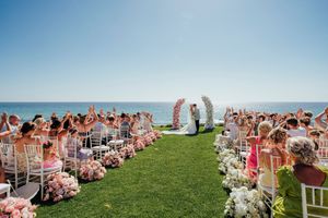 Panoramic view of outdoor wedding ceremony guests seated in white chairs on a grassy hilltop overlooking the ocean.