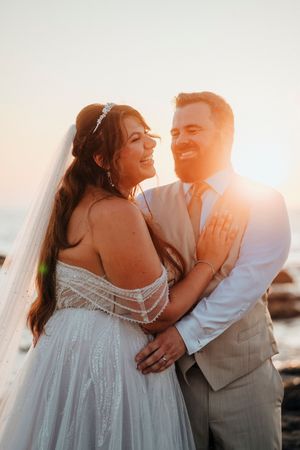 A couple shares a romantic sunset moment at their beach wedding wearing white dress and gray suit.