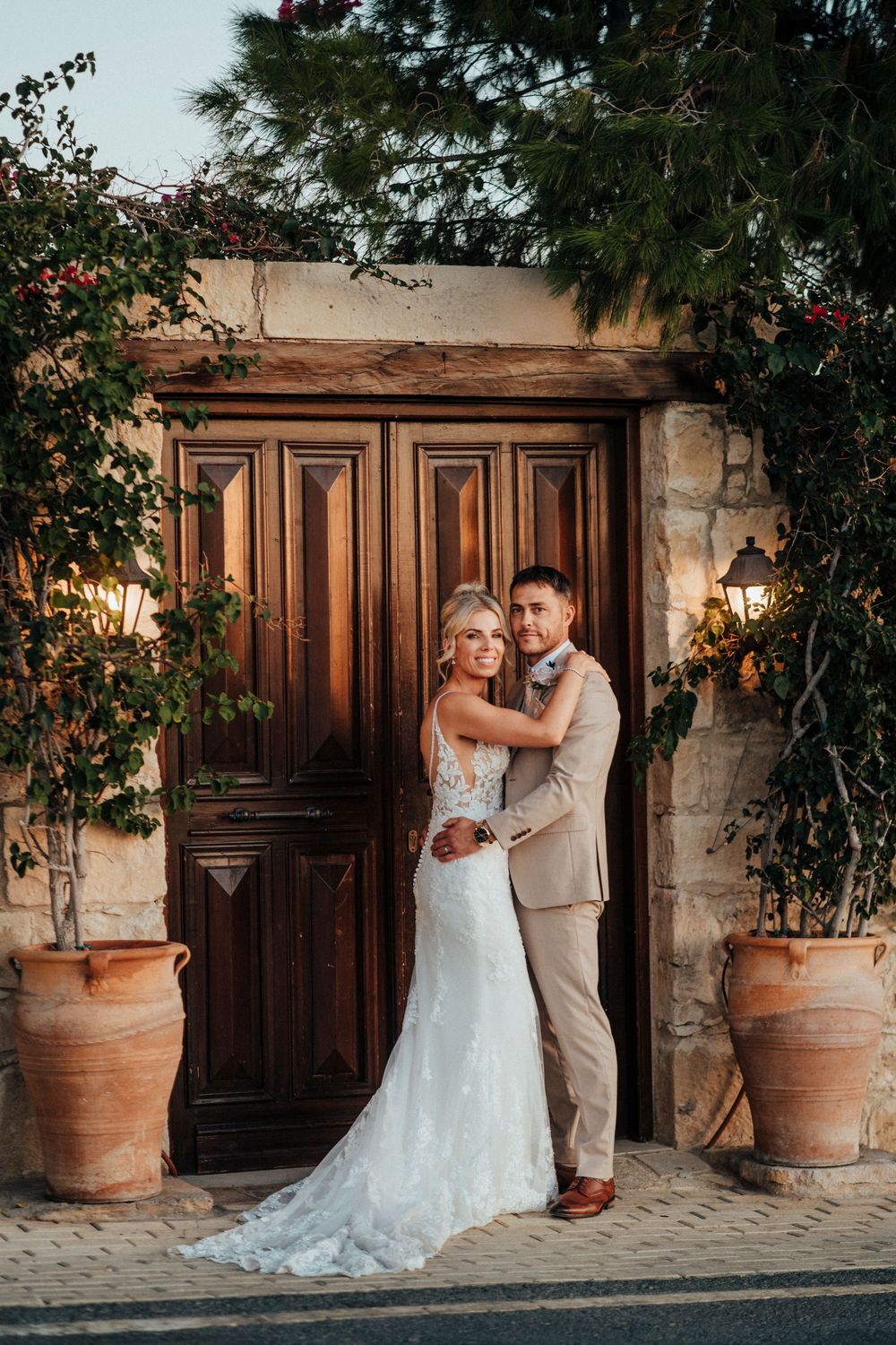 Romantic wedding couple poses in front of rustic wooden doors with terracotta planters at sunset.