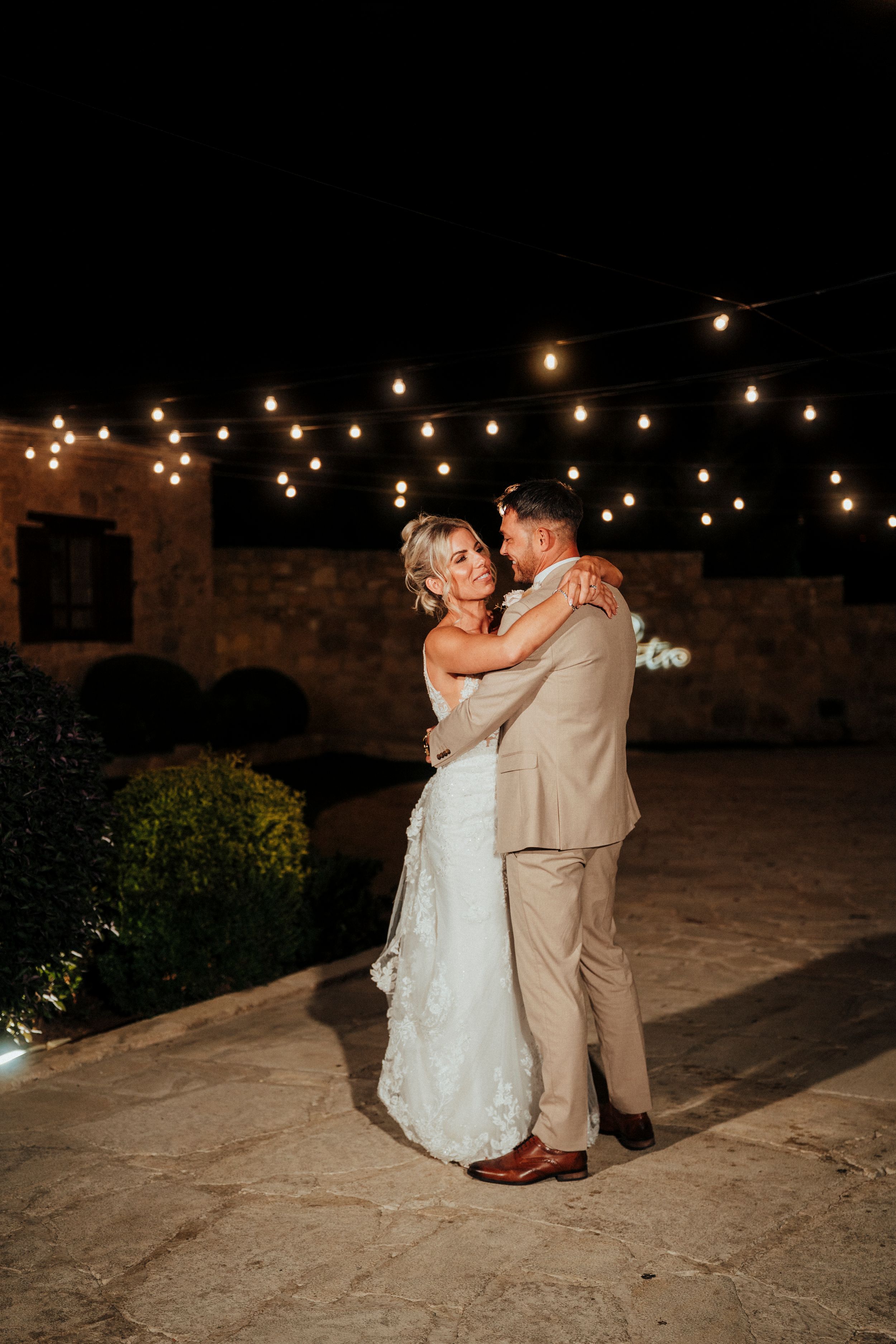 Newlyweds share a romantic dance under string lights at a stone venue during their evening wedding reception.