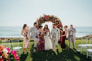 A wedding celebration on a coastal lawn with a floral arch and white chairs overlooking the ocean.