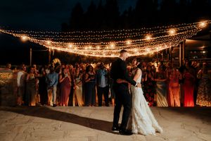 Wedding reception dance floor illuminated by string lights with guests gathered around under the evening sky.