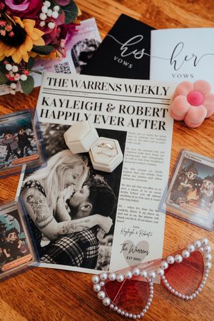 Wedding stationery and decorative items including invitations, newspaper-style programs, and pearl heart decorations on wooden surface.
