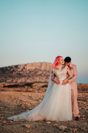 A couple in wedding attire poses together in a desert landscape at sunset, creating romantic silhouettes against rocky terrain.