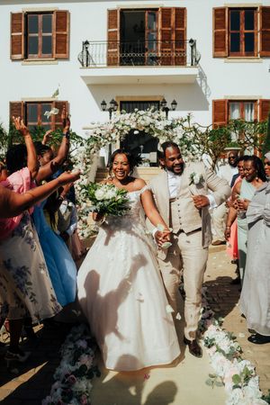 Wedding party celebrates joyfully with dancing and raised arms against white building exterior with wooden shutters.