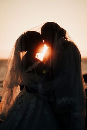 Silhouetted wedding couple embracing against warm sunset sky with flowing veil creating dramatic backlit effect.
