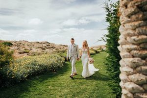 A couple walks hand in hand through a scenic meadow with wildflowers and mountains in the background.
