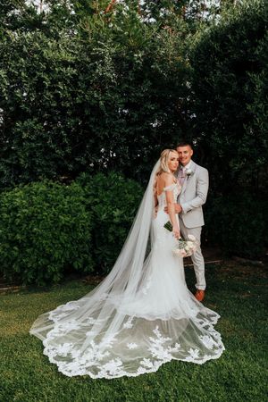 A couple in wedding attire embraces in a garden setting with a stunning lace veil trailing behind them.