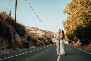 A couple stands in the middle of a winding mountain road surrounded by golden sunlit trees at sunset.