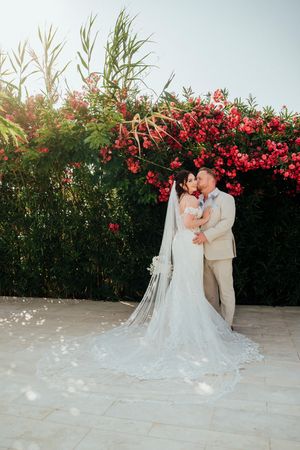 A romantic wedding couple shares a tender moment against a backdrop of vibrant red flowering bushes at sunset.