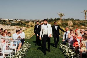 Wedding party walks down outdoor aisle decorated with white flowers and chairs on grassy venue with palm trees.