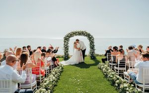 Outdoor wedding ceremony on a grassy cliff overlooking the ocean with floral arch and white chairs for guests.