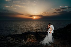 Romantic wedding couple embraces on a dramatic clifftop overlooking the ocean at sunset.