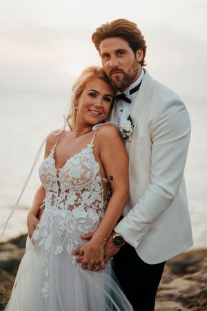 Romantic coastal wedding portraits at sunset with couple in white formal attire embracing on rocky shore.