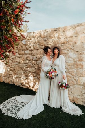 Two people in elegant white wedding dresses pose together against a rustic stone wall with vibrant floral arrangements.