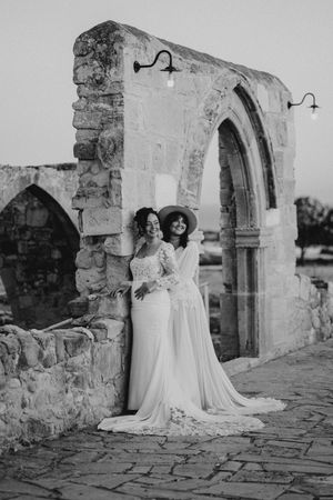 Two brides in elegant white wedding gowns pose together at a historic stone archway in black and white photography.