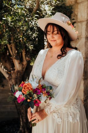Bride in vintage-inspired lace wedding dress with long sleeves holds vibrant coral and pink floral bouquet in garden setting.