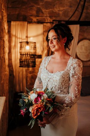 A bride in a long-sleeve lace wedding dress holds a vibrant autumn bouquet in a rustic venue with warm lighting.