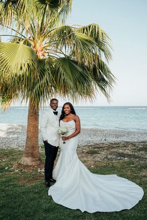 A couple in wedding attire poses romantically under palm trees by the ocean at sunset.