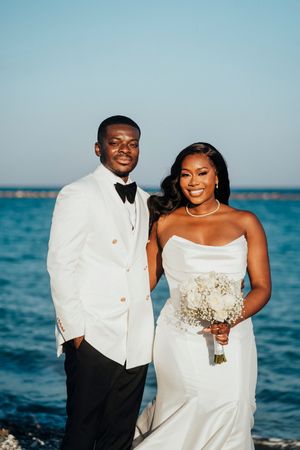 A couple in elegant white wedding attire poses for romantic photos against a beautiful blue ocean backdrop.