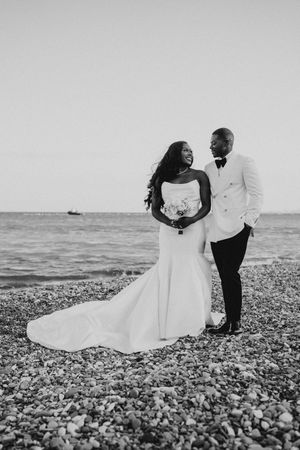 A romantic black and white wedding photo series showing a couple embracing on a pebble beach by the ocean.