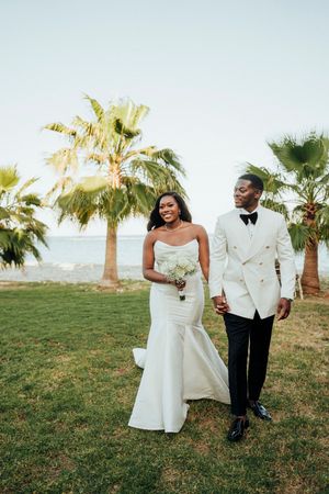 A couple in elegant white wedding attire walk together on a grassy hill with palm trees in the background.
