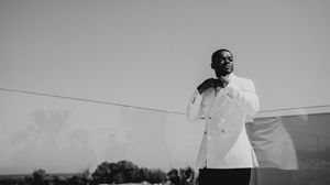 A person in a white blazer stands outdoors against an overcast sky while holding a microphone.