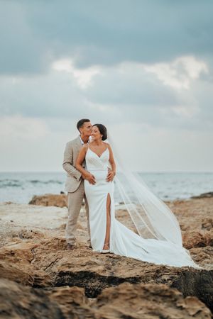 A couple in wedding attire share an intimate moment on rocky coastal cliffs with ocean waves and cloudy skies in background.