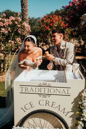 Wedding couple enjoying ice cream from a traditional ice cream cart surrounded by blooming flowers and palm trees.