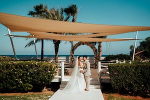 A beautiful outdoor wedding ceremony setup with beige canopies and palm trees against a bright blue sky.