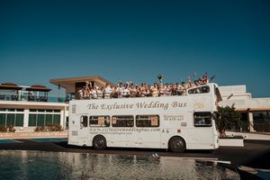 A white wedding bus transports guests along a scenic coastal road with blue waters and clear skies in view.